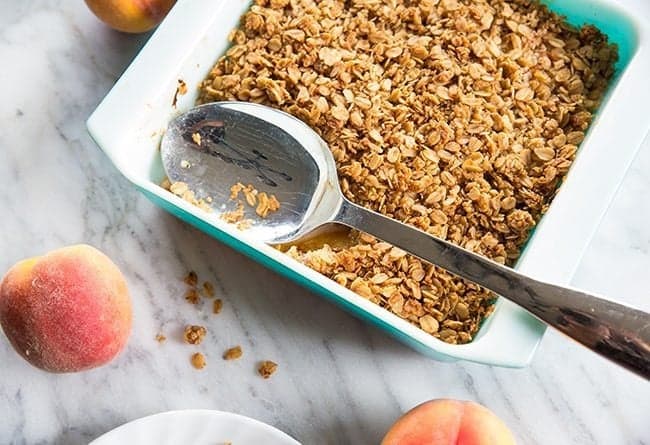 Peach Crisp in a white plate with spoon, peach fruit on a marble background