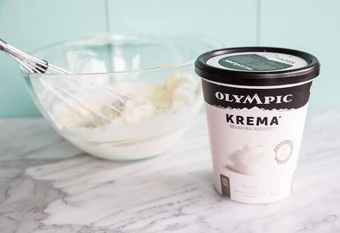 a cup of Olympic Krema Greek Style Thick & Rich Yogurt and a large transparent bowl with a whisk and yogurt