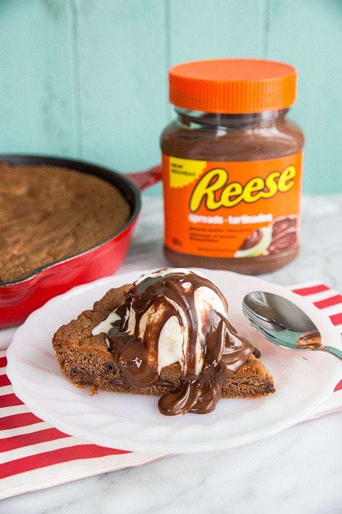 butterscotch ripple ice cream with chocolate sauce on top of skillet cookie slice, reese's spread and red skillet on background