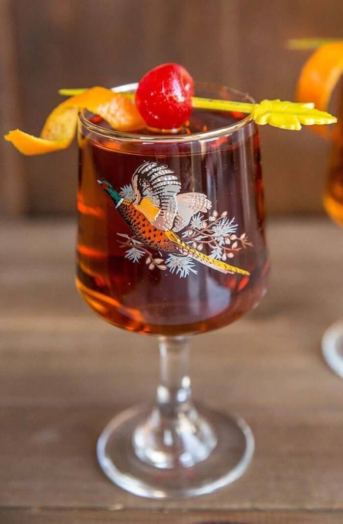 Close up of Manhattan cocktail drink in vintage pheasant glass with cherry and orange garnish