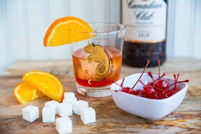  a glass of Don Draper Classic Old Fashioned Cocktail with a slice of orange, some fresh cherries in a small dip bowl