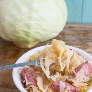 Kielbasa Kapusta in a small white bowl with fork in it, whole cabbage on background