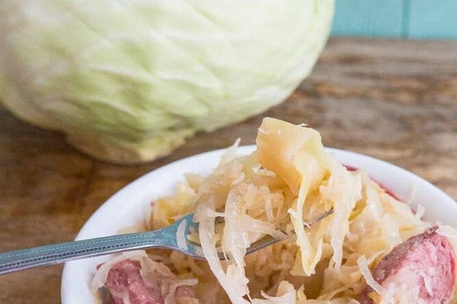 Kielbasa Kapusta in a small white bowl with fork in it, whole cabbage on background