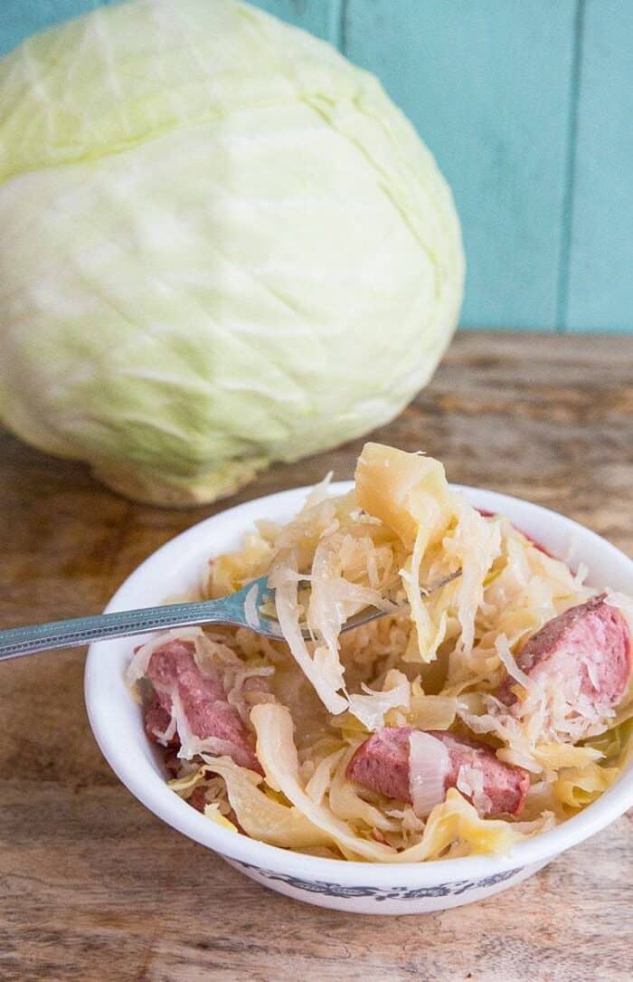 Kielbasa Kapusta in a small white bowl with fork in it, whole cabbage on background