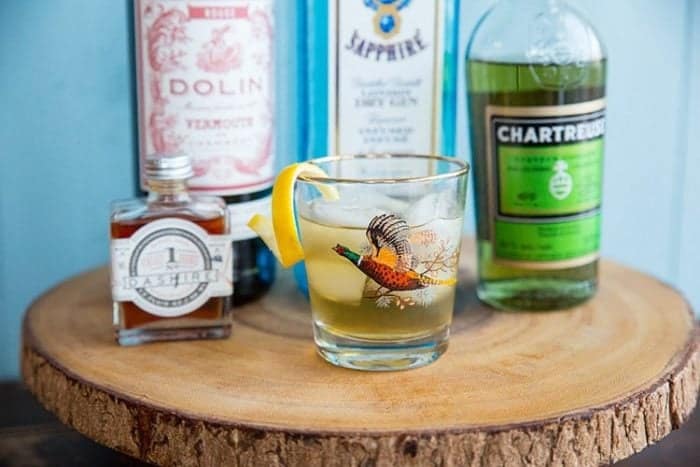 A glass of Bijou Cocktail in a round wood stand, bottles of dry Gin, Green Chartreuse, sweet Vermouth and orange bitters at the back