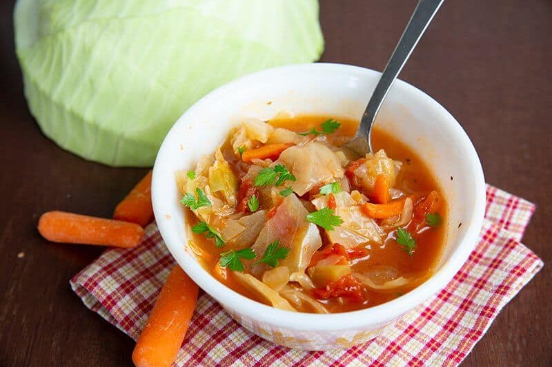 Checkered red kitchen cloth underneath cabbage soup in a bowl with a cabbage and carrots.