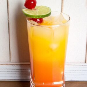 Close up of a glass with tequila sunrise garnish with a slice of lime and 2 maraschino cherries on pick