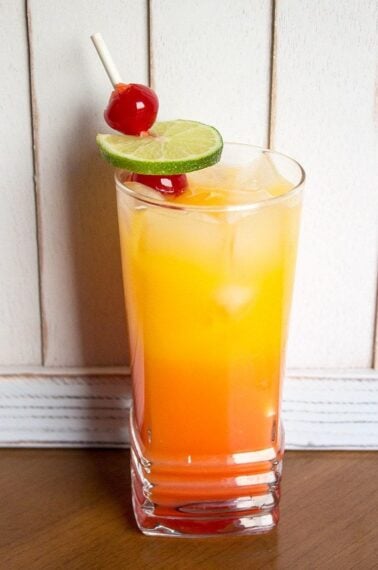 Close up of a glass with tequila sunrise garnish with a slice of lime and 2 maraschino cherries on pick