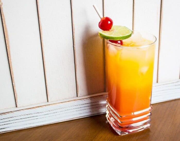 A glass with tequila sunrise garnish with a slice of lime, and 2 maraschino cherries on pick