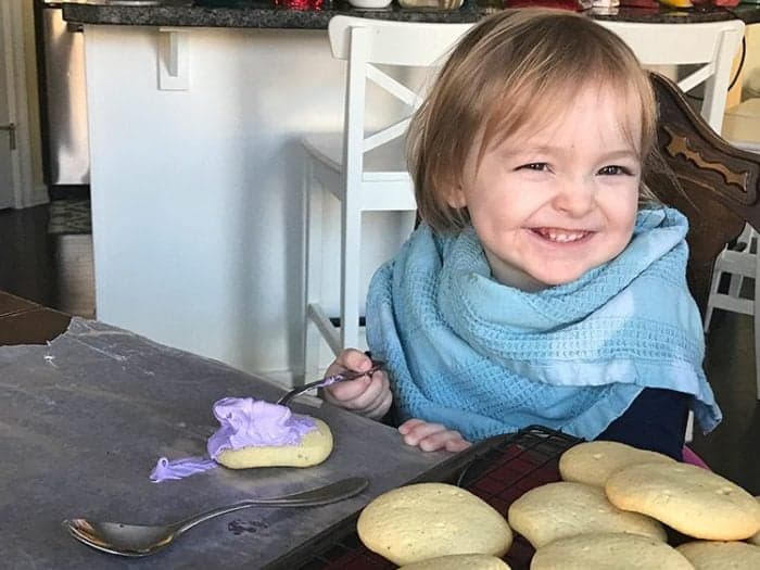 little girl holding a spoon with buttercream frosting, some cut-out sugar cookies in front of her