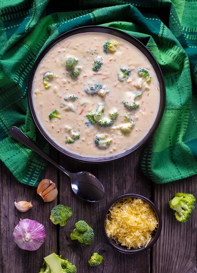 green tablecloth underneath a bowl of Broccoli Cheese Soup with spoon, some of its ingredients in a wooden background