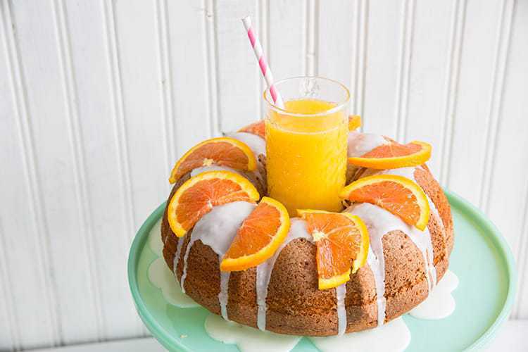 Harvey Wallbanger Cake with Boozy Glaze and slices of oranges on top, a glass of orange juice at the center of bundt cake