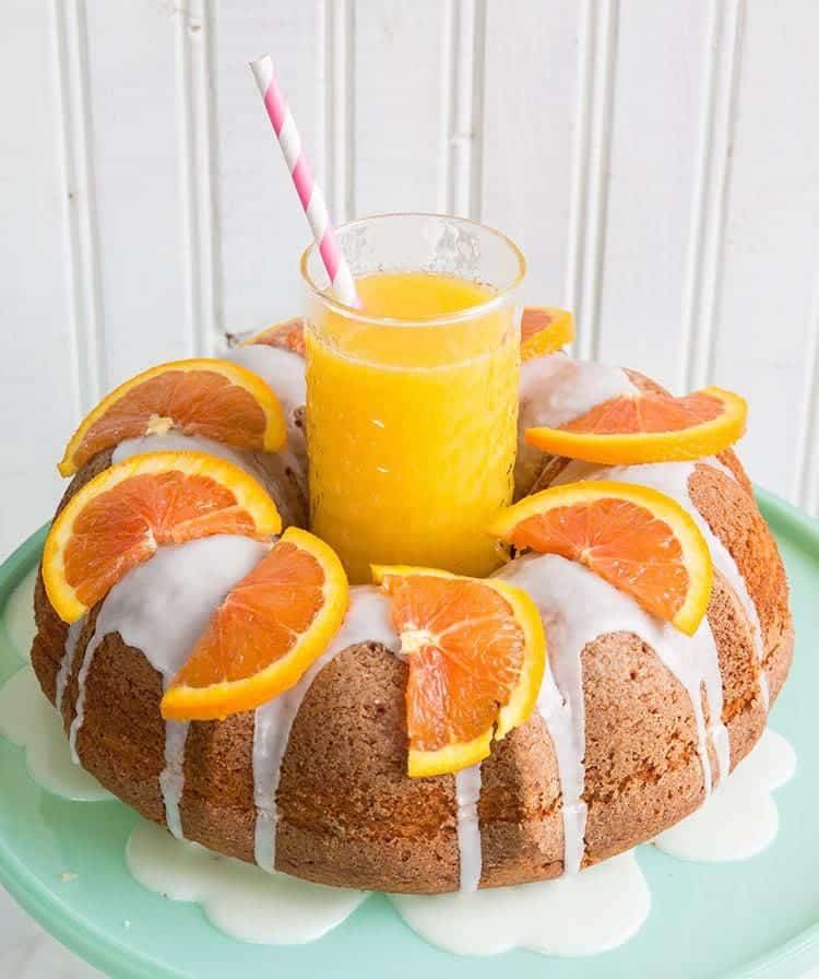 Close up Harvey Wallbanger Cake with Boozy Glaze and slices of oranges on top, a glass of orange juice at the center of bundt cake
