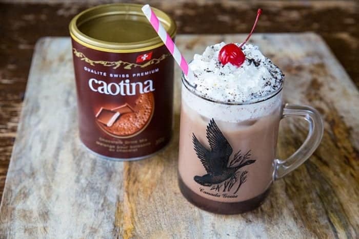 A glass of Spiked Chocolate Iced Coffee topped with whipped cream and cherry, a can of Caotina beside it on a wooden background