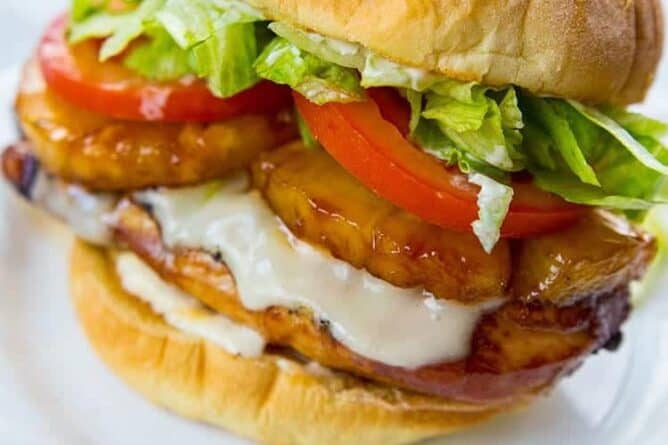 white plate with a Teriyaki Chicken Burger loaded with fillet, pineapple slices, Swiss cheese, tomato slices and shredded lettuce