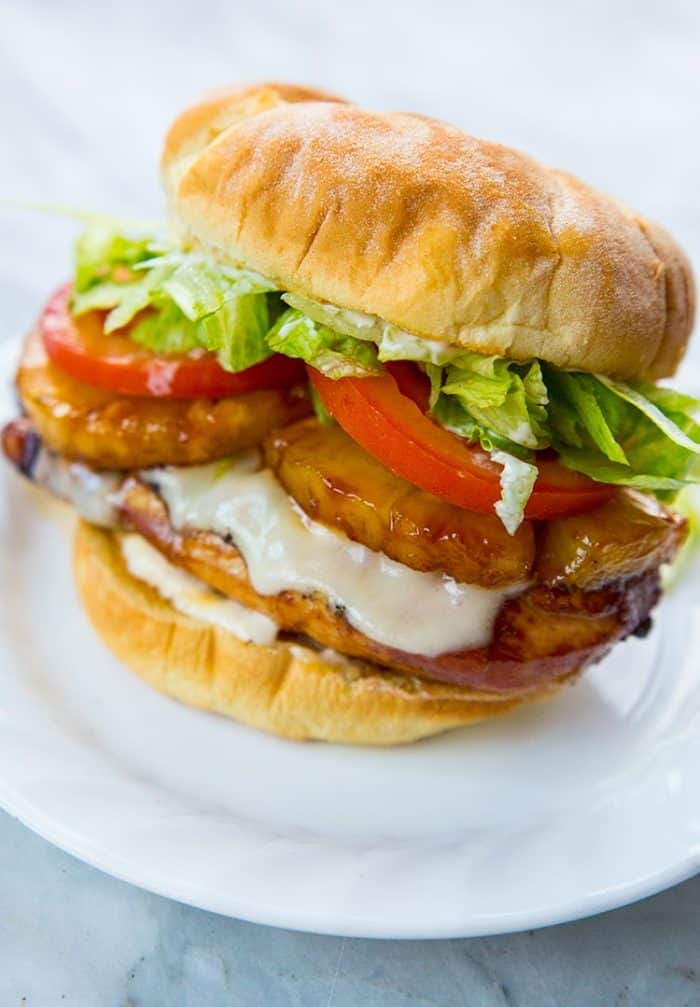white plate with a Teriyaki Chicken Burger loaded with fillet, pineapple slices, Swiss cheese, tomato slices and shredded lettuce
