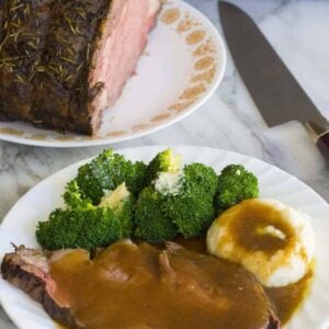 Prime Rib roast plated with broccoli and mashed potatoes topped with gravy. A knife and a plate with whole prime rib roast on background