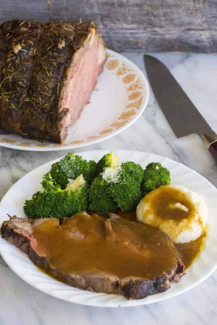 Prime Rib roast plated with broccoli and mashed potatoes topped with gravy. A knife and a plate with whole prime rib roast on background