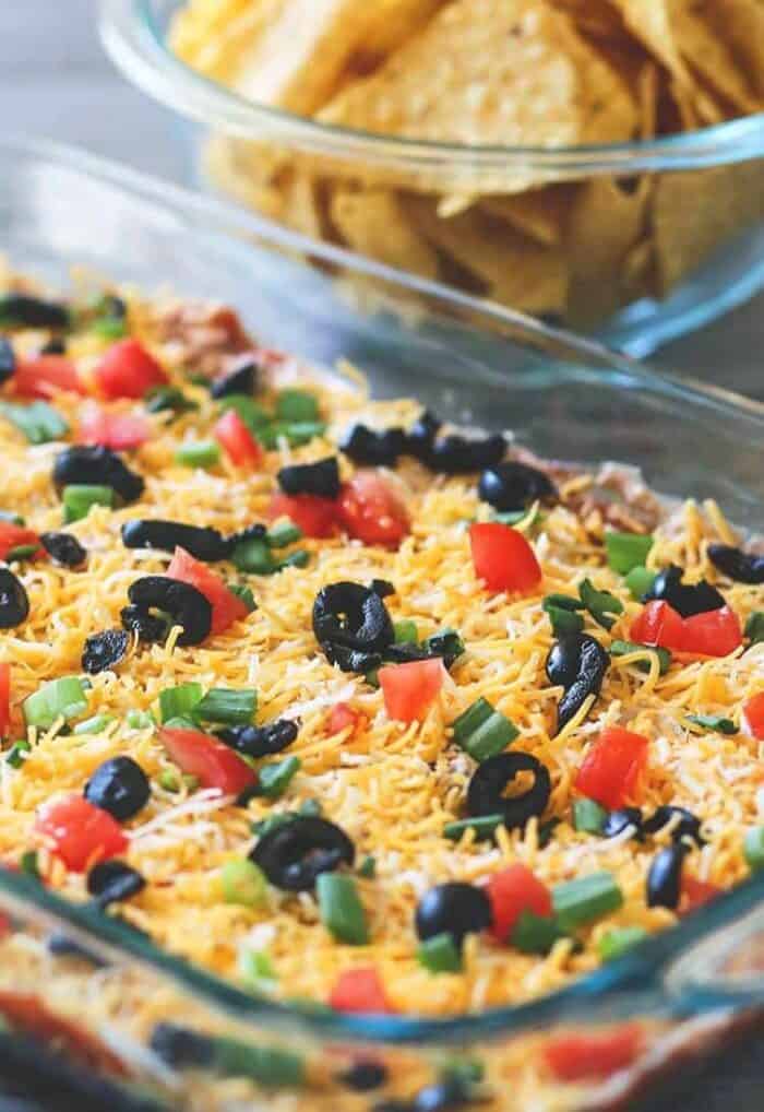 Pyrex baking dish with Tex-Mex 7 Layer Dip topped with tomatoes, green onions, shredded cheese and black olives