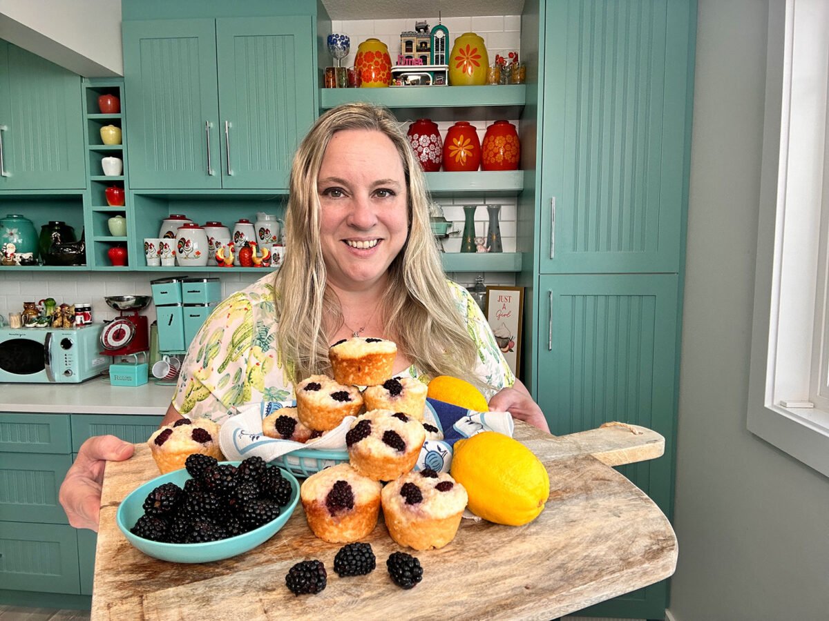 Karlynn holding a wooden cutting board with a bowl of blackberries and a stack of muffins and lemons on it. 