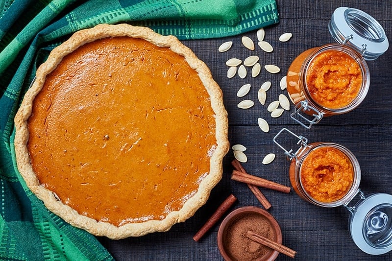green table cloth underneath a pie pan with Pumpkin Pie, some pumpkin seeds cup of ground cinnamon sticks and canned pumpkin in a container beside it