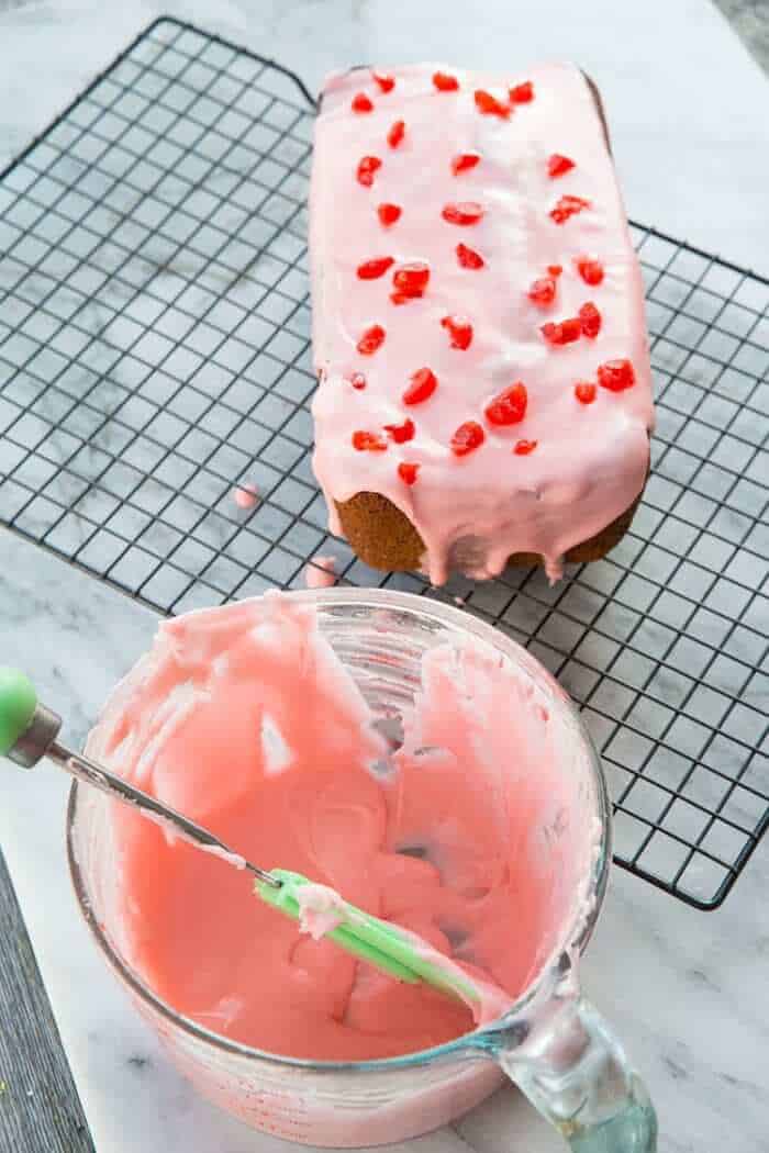 sliced cherry poppy seed loaf cake with glaze on wire baking rack, a bowl with cherry icing glaze on its side