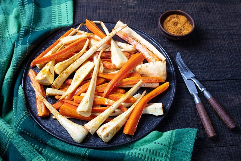 green tablecloth underneath a black serving plate with roasted parsnips and carrots, Cajun seasoning bread knife and fork on its side