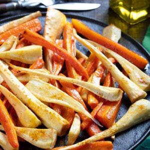 close up black serving plate with roasted parsnips and carrots, Cajun seasoning, olive oil, bread knife and fork on its side