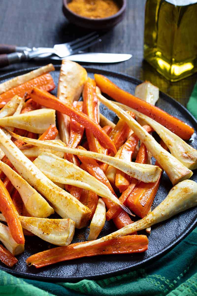 close up black serving plate with roasted parsnips and carrots, Cajun seasoning, olive oil, bread knife and fork on its side