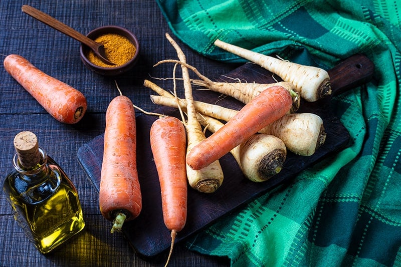 Parsnips and Carrots on a wooden cutting board with green tablecloth underneath, olive oil and cajun seasoning on its side