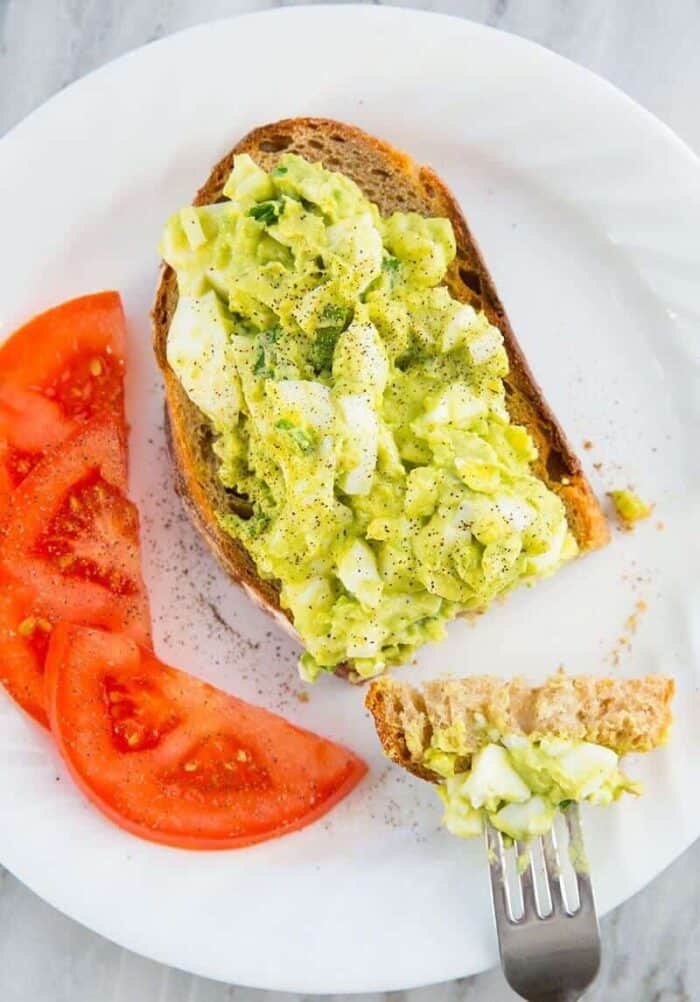 close up of white plate with slices of red tomatoes and open faced toasted bread topped with Avocado Egg Salad