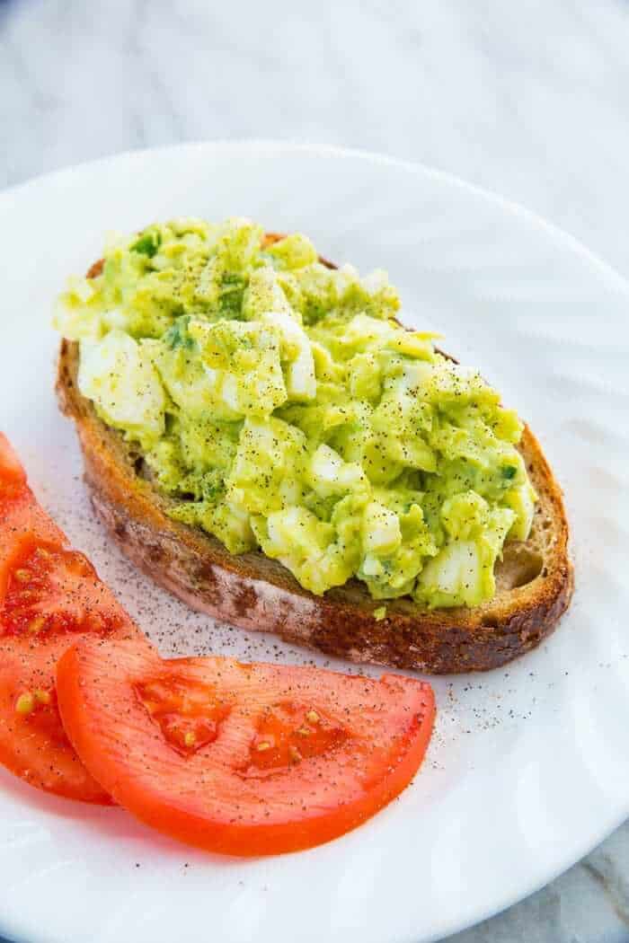 crusty toasted piece of sourdough with Avocado Egg Salad on top, slices of red tomatoes beside it with salt and pepper