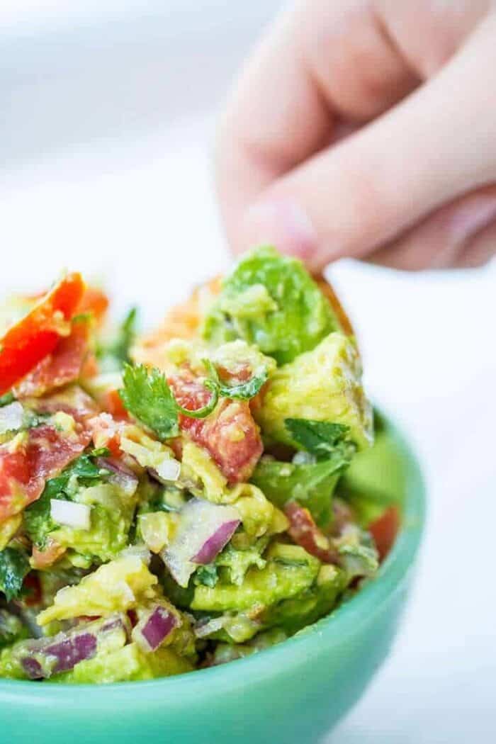 close up hand dipping chips into guacamole in a blue bowl