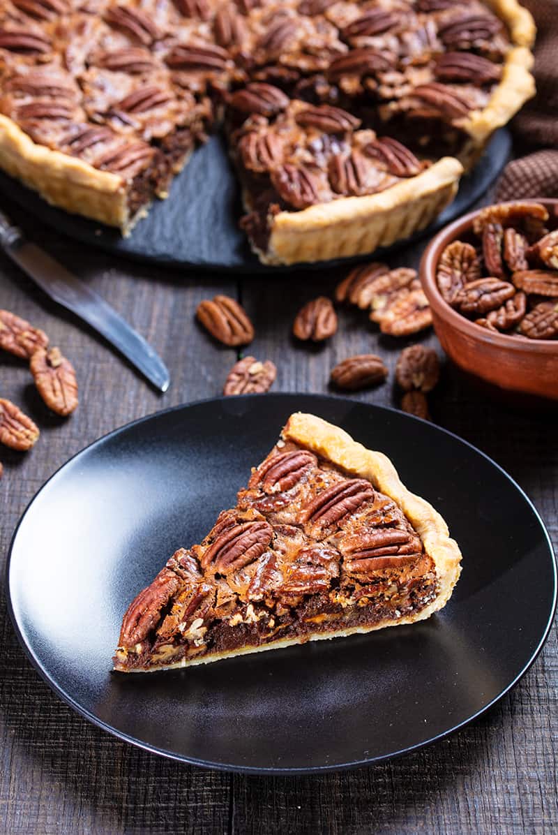 a slice of Chocolate Pecan Pie on a black medium serving plate, a cup of pecans and the remaining part of the pie on its background