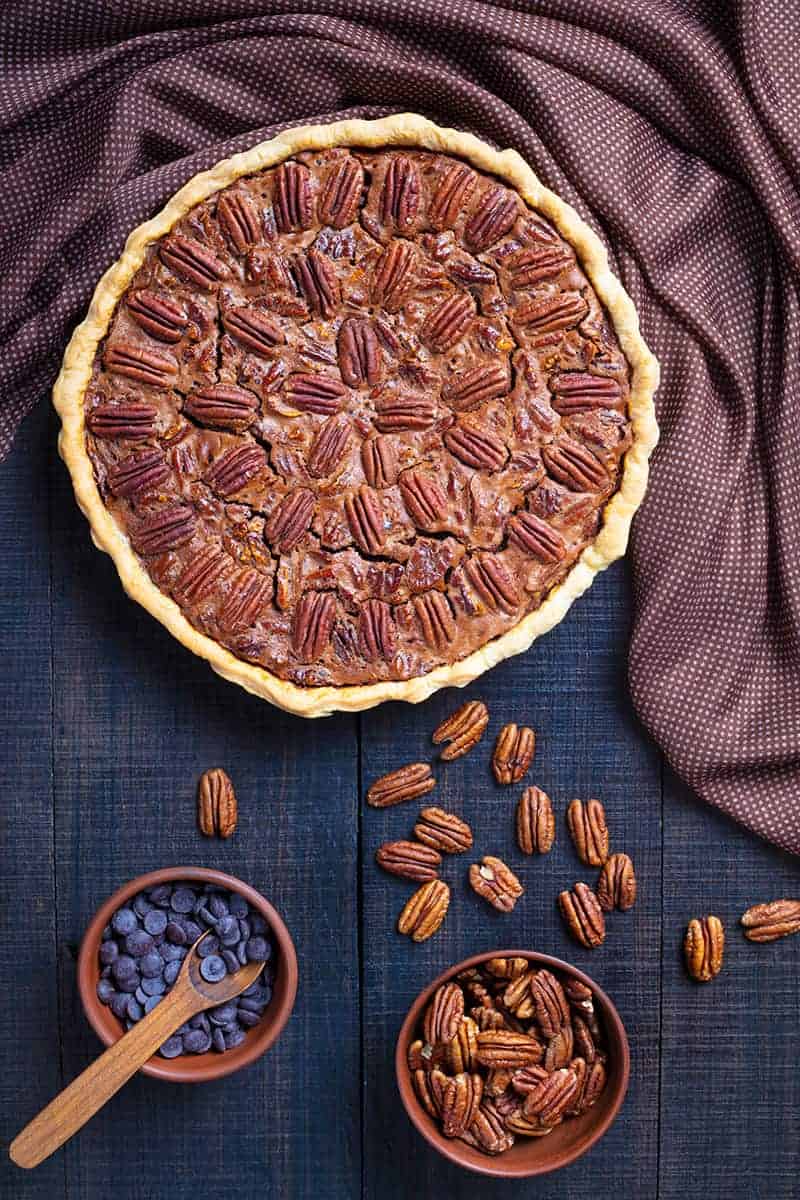 brown polka dot tablecloth on dark wood background, whole Chocolate Pecan Pie with a cup of whole pecan and a cup of chocolate chips beside it