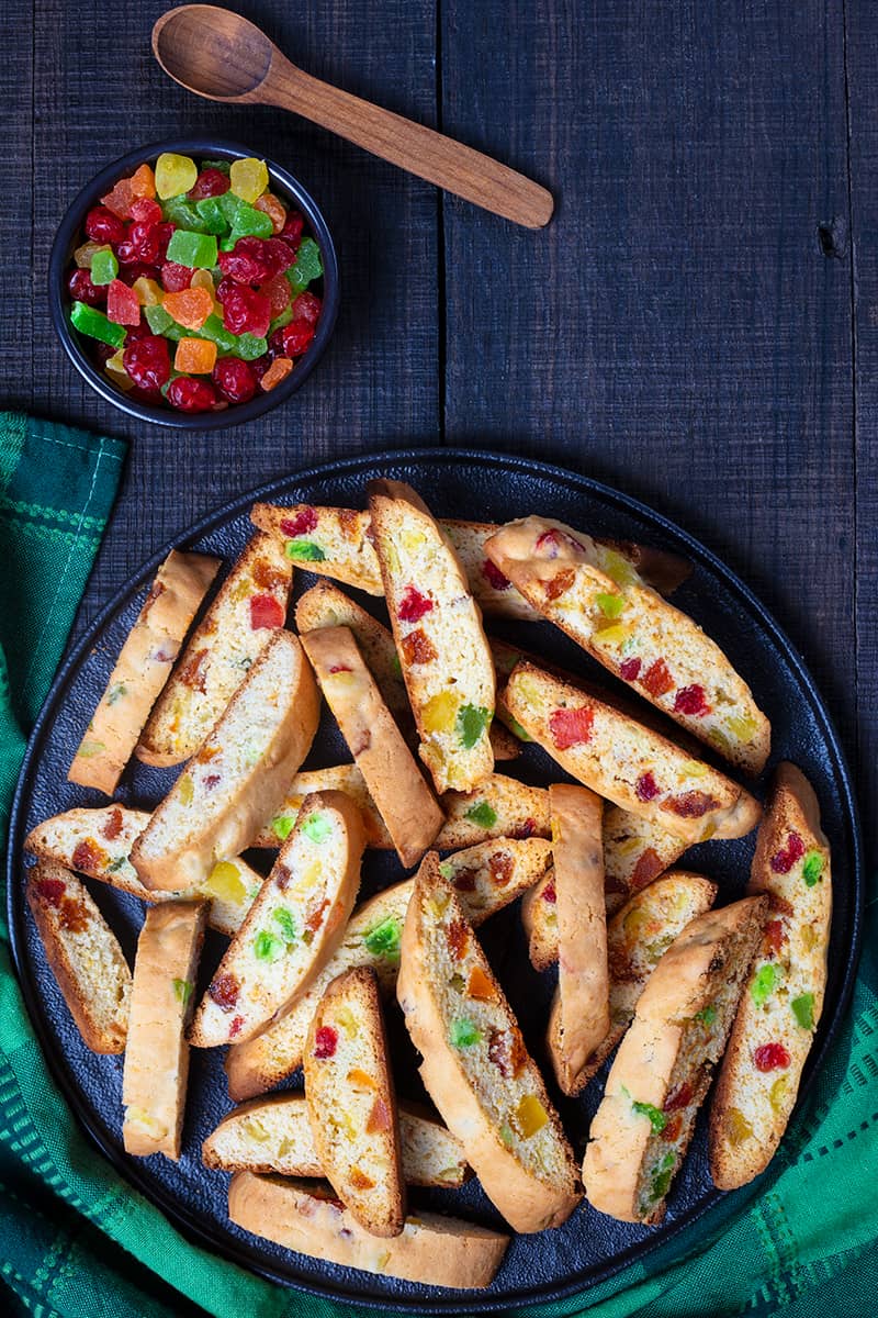 green tablecloth underneath a large black serving plate with slices of toasted Christmas Biscotti, a cup of candied fruits and a wooden spoon beside it