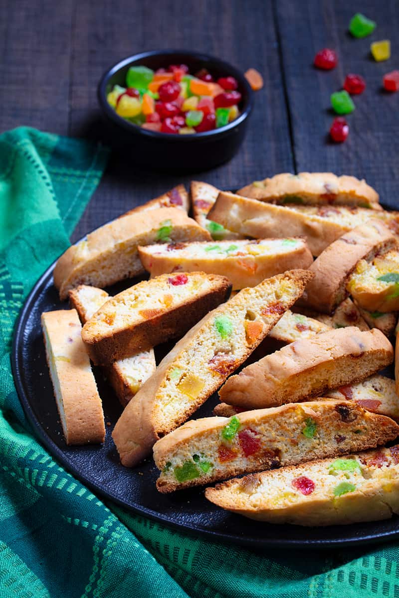 close up green tablecloth underneath a large black serving plate with slices of toasted Christmas Biscotti, a cup of candied fruits beside it