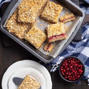 blue checkered tablecloth underneath a baking pan lined with parchment paper with Cranberry Oatmeal Bars, cranberries and white plate with a piece of Cranberry Oatmeal Bar on its side