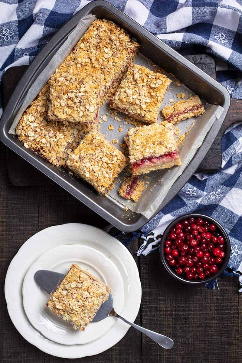 blue checkered tablecloth underneath a baking pan lined with parchment paper with Cranberry Oatmeal Bars, cranberries and white plate with a piece of Cranberry Oatmeal Bar on its side