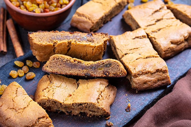 slices of soft and chewy Molasses Bars (Hermit Bars) in a wooden cutting board, a cup of raisins and cinnamon stick on background