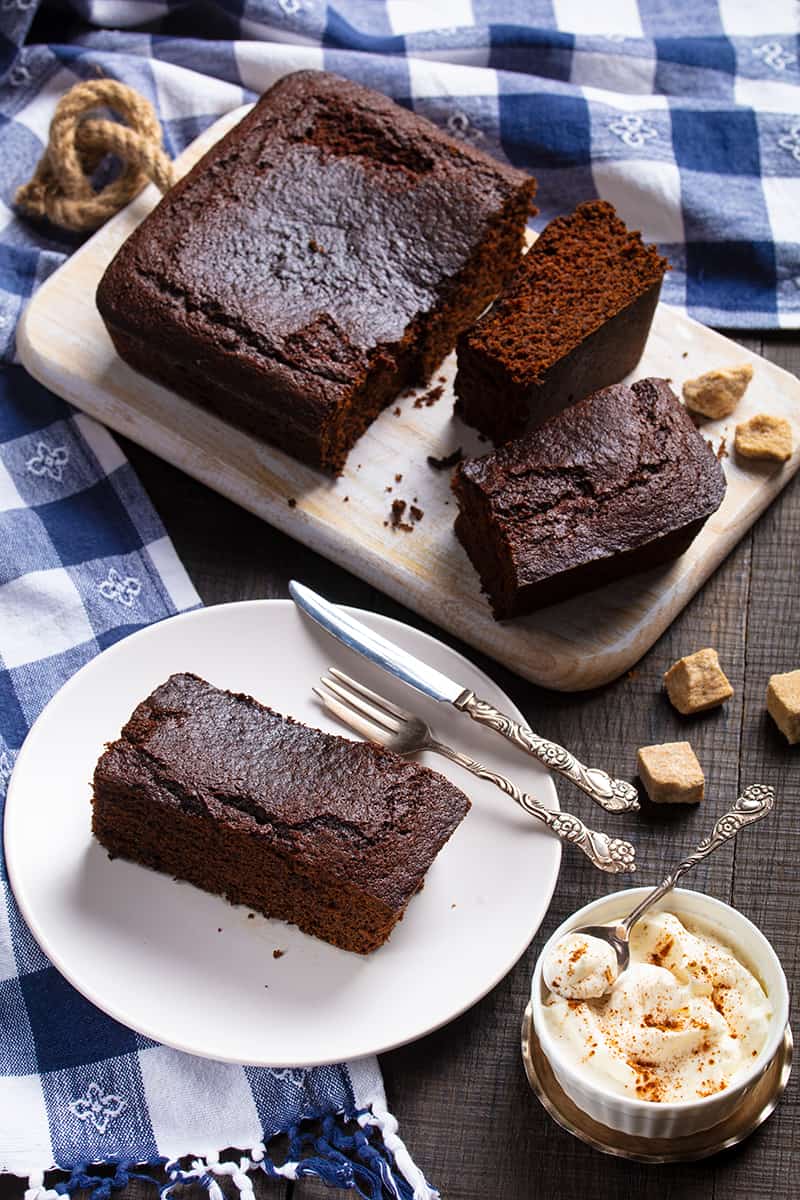 close up Old Fashioned Gingerbread slices on wooden cutting board and white plate with fork and bread knife, a cup of butter cream icing on its side