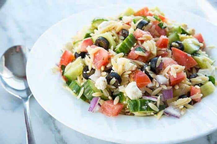 Greek Orzo Salad in a white bowl with a spoon beside on marble background 