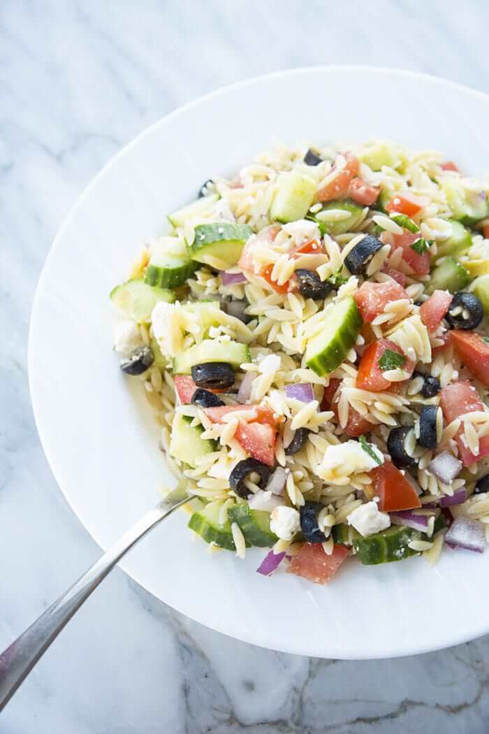 Greek Orzo Salad- tomatoes, orzo, black olives feta and purple onions in a white bowl