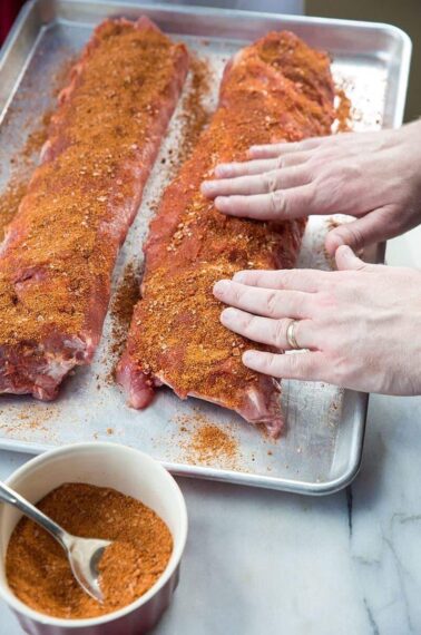 Raw pork ribs being prepared with rib rub on a baking sheet
