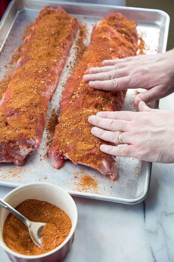 Raw pork ribs being prepared with rib rub on a baking sheet