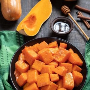 green tablecloth underneath black serving plate with Honey & Cinnamon Roasted Butternut Squash, fresh squash honey and cinnamon sticks on side