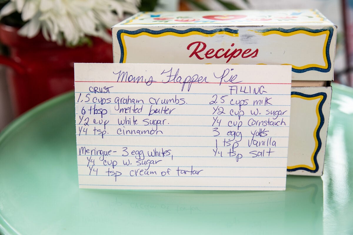 A handwritten recipe card for “Mom’s Flapper Pie” sits on a pastel green surface, in front of a vintage recipe box decorated with a red and yellow pattern. This classic Flapper Pie recipe is surrounded by an inviting nostalgia, with flowers slightly visible in the background.