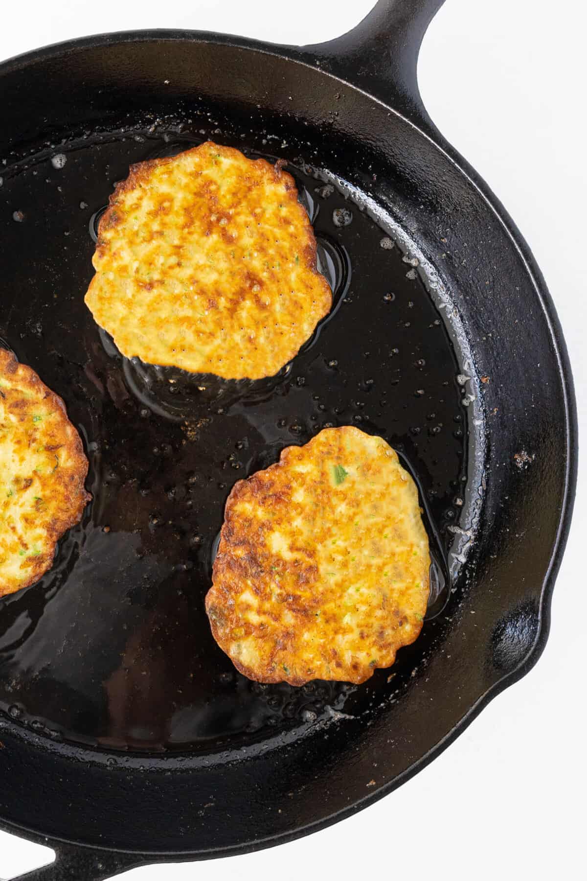 cooking three zucchini fritters in a cast iron pan