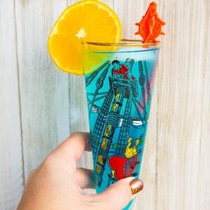 woman's hand holding a blue AMF cocktail drink garnished with an orange slice that's in a vintage pirates motif glass