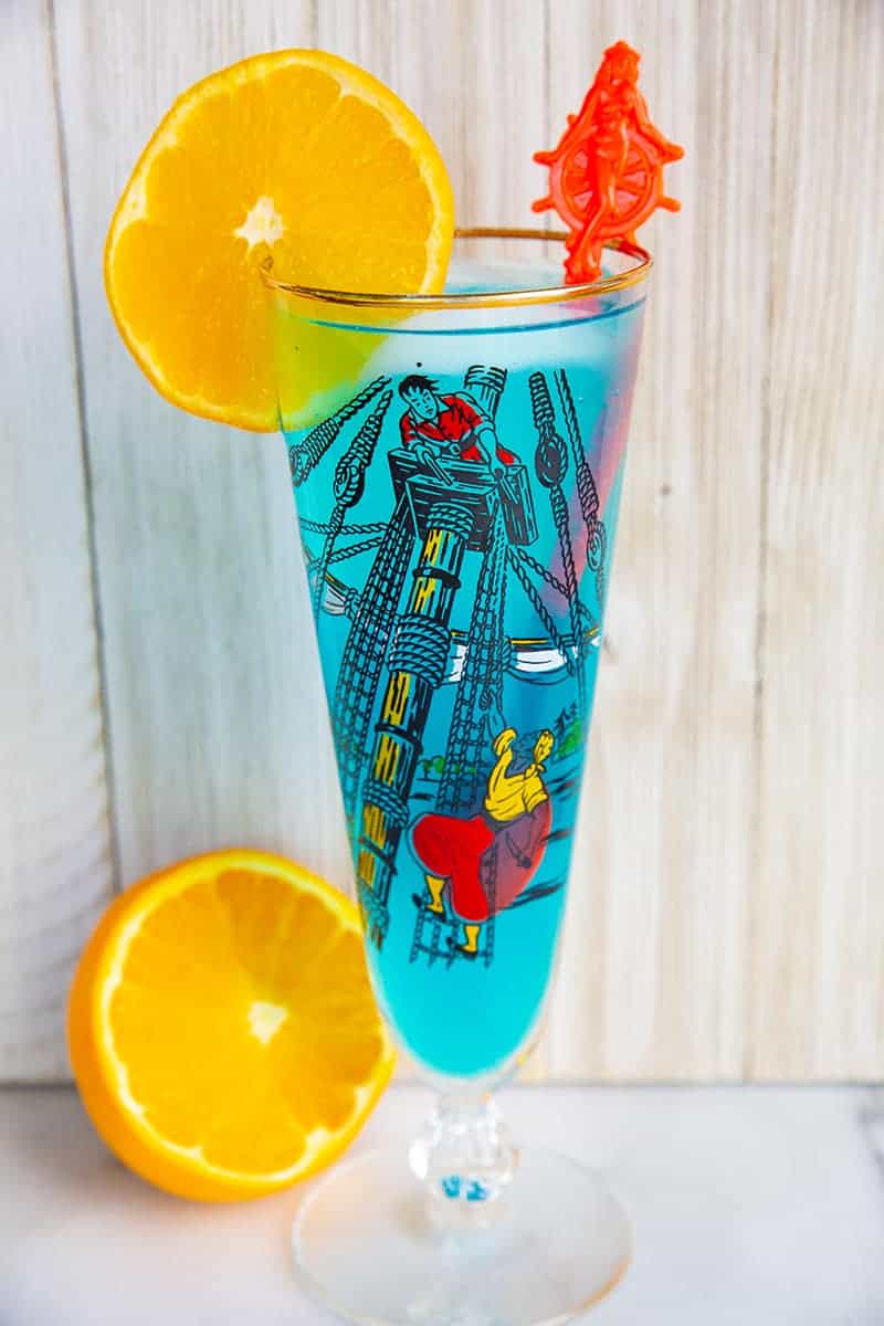A blue AMF cocktail drink garnished with an orange slice that's in a vintage pirates motif glass